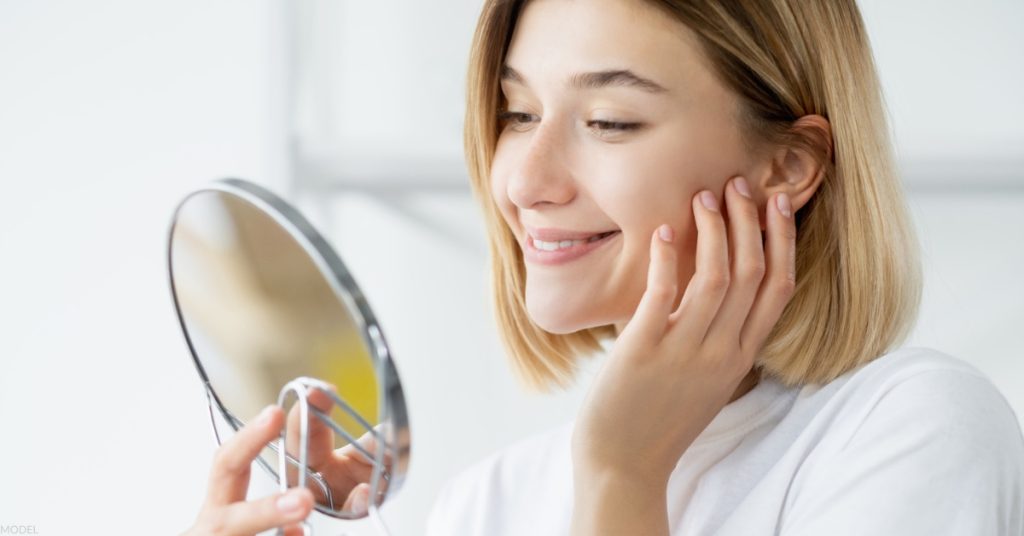 Woman admiring her appearance in the mirror after skincare treatment (Model).