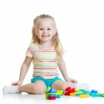 A smiling young blond girl sits on the floor with colorful blocks in front of her.