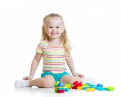 A smiling young blond girl sits on the floor with colorful blocks in front of her.