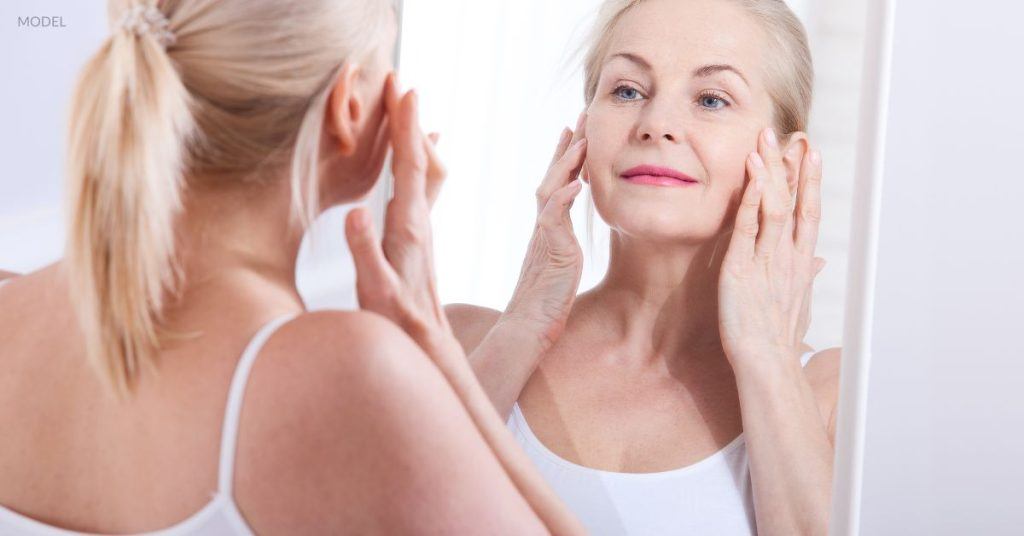 Mature woman with a youthful-looking face (model) holds hands up to her face while looking in the mirror.