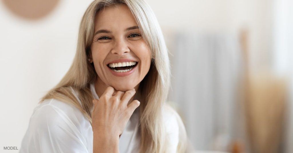 Woman with clear youthful looking skin (model) smiling with a hand rested under her chin.