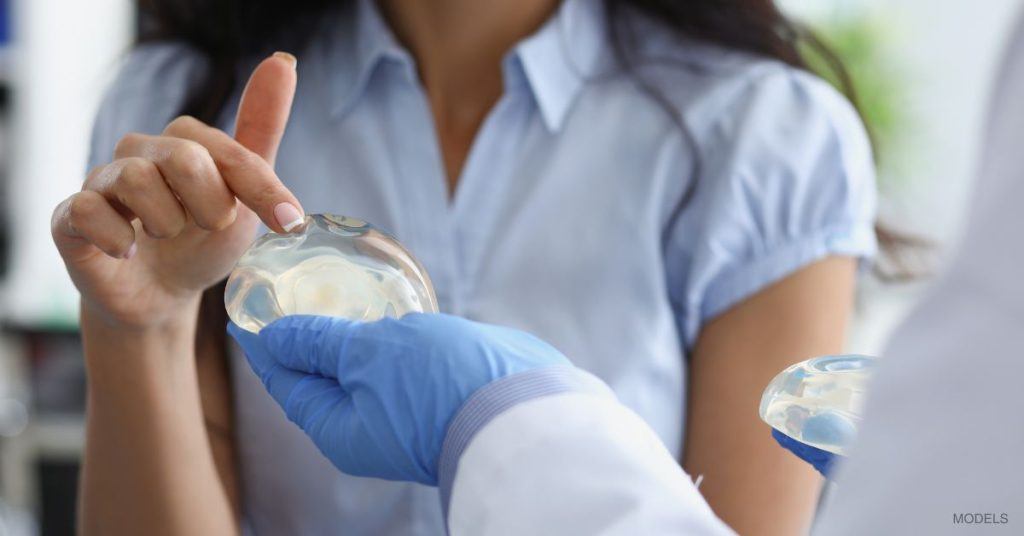 Woman and doctor (models) meet in an office for a breast implant consultation. The woman is reaching out to feel one of the available implants.