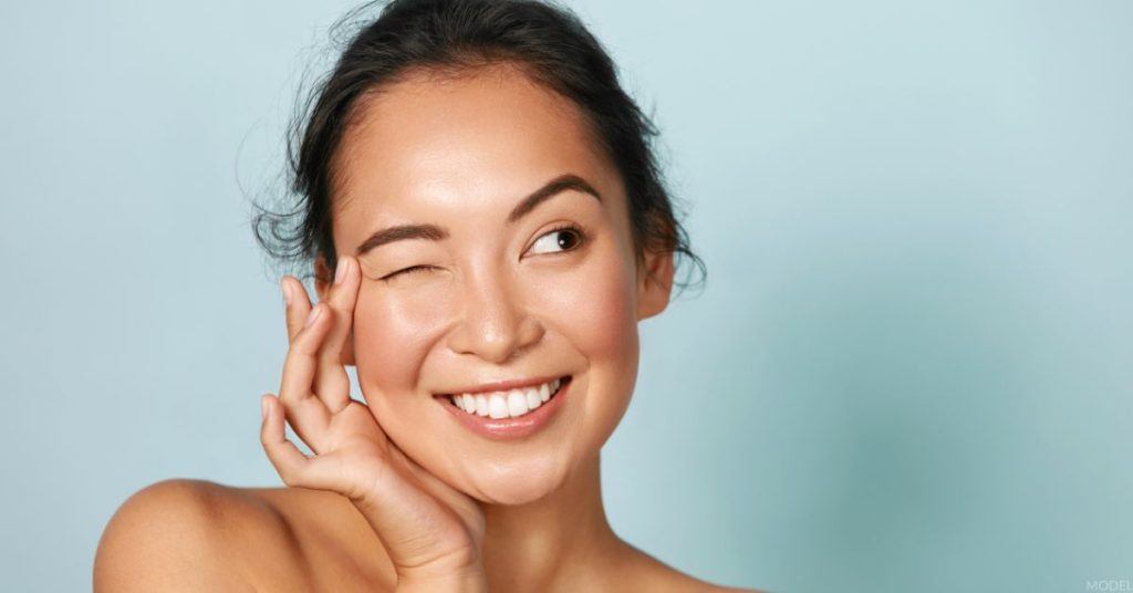 Woman smiling and winking, gently touching the area around her eye. (MODEL)